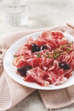 Freshly cut dried meat, served with olives and herbs, on a white plate, light background, no people