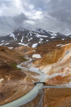 Tourist on bridge over steaming stream between colourful rhyolite mountains with snowfields,