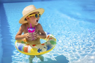 Playful caucasian male kid in sun hat and sunglasses on a pool with a glass of water standing