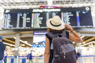 A woman wearing a straw hat and a backpack is looking at a large airport screen. She is waiting for