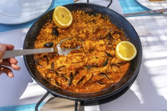 Close up of a spoon scooping up seafood paella from a traditional paella pan, garnished with lemon
