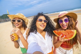 Three women are smiling and holding watermelons on a beach. They are wearing straw hats and