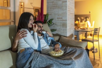 Young couple enjoying a movie night at home, eating popcorn from a bowl and relaxing on the sofa