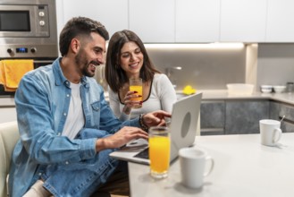 Happy couple using laptop and drinking orange juice while having breakfast in modern kitchen