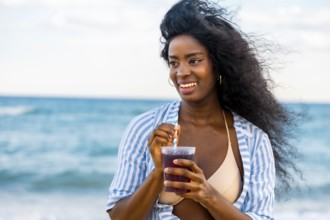 Smiling african american woman enjoying a refreshing drink on a sunny beach, with blue sea in the