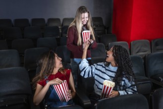 Three female friends are laughing and sharing popcorn while watching a movie in a cinema, enjoying