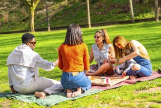Group of diverse friends enjoying pizza and laughter during a sunny picnic in a vibrant park,