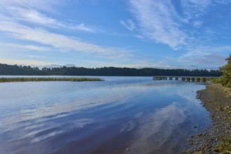 A jetty and shingle beach stretch along the shore of a tranquil lake under blue skies, Lake