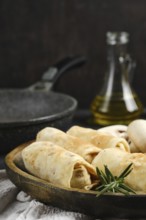 Close-up view of crepes with mushrooms on a wooden plate. In the background, an oil bottle and a