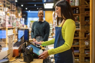 Smiling saleswoman wearing apron using touchscreen cash register while customer waiting at checkout
