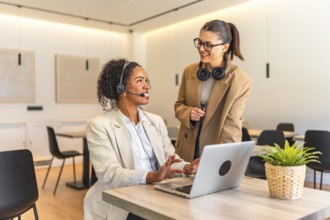 Two smiling female customer service operators working together on a laptop in a modern office