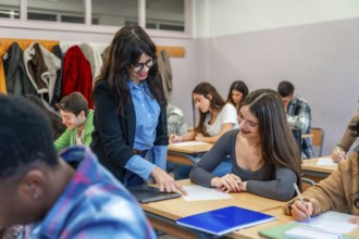 Female professor assisting a smiling student with her exam paper in a classroom full of diverse