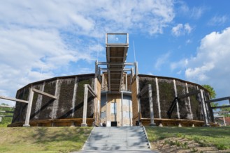 Round building made of wood and metal with stairs, blue sky and green landscape in the background,