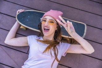 Happy young woman lying on a wooden floor with her skateboard showing peace sign, wearing pink