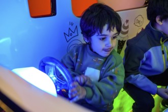 Young boy playing with toy car steering wheel, enjoying entertainment in colorful amusement park