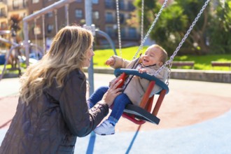 Happy mother pushing her laughing baby son on a swing, enjoying a sunny autumn day at the
