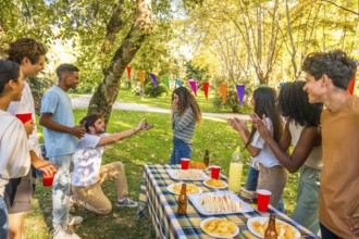 Caucasian young man kneeling to propose marriage to woman during picnic with multi-ethnic friends