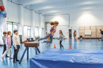 Elementary school girl practicing gymnastics, jumping over a pommel horse in the school gym watched