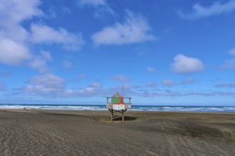 A rescue tower stands alone on a wide sandy beach under a blue sky, Bethells Beach, Te Henga,