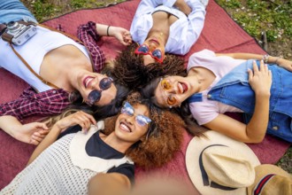 Four cheerful young women with sunglasses and hats are lying in circle on a blanket at a park,