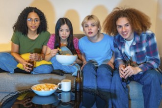 Four young friends enjoying a night in, watching television, eating snacks and drinking beer,