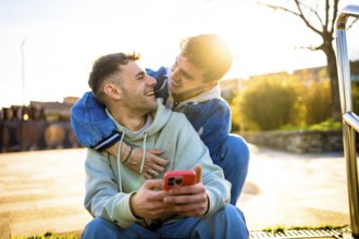 Two men hugging and laughing while using a smartphone outdoors, enjoying a sunny day together