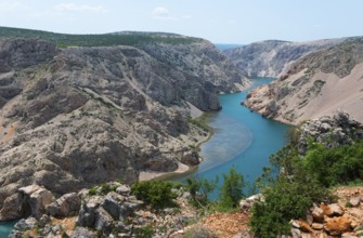 A deep gorge with a turquoise-blue river, surrounded by barren rocks, under a summer sky,