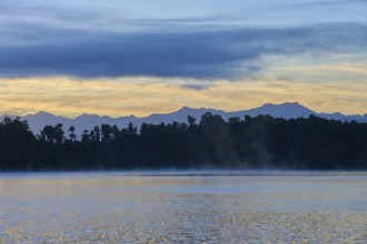 A hazy lake at sunrise with a mountainous silhouette and blue surroundings, Lake Mahinapua,