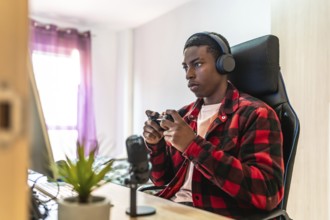 Young man playing videogames on a desktop computer using a controller, wearing headphones and using
