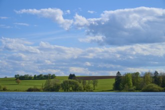 Ober- Moosersee surrounded by green fields and trees under a partly cloudy blue sky, spring,