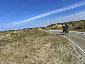 Cyclist on a wide cycle path through a dune landscape under a blue sky with clouds, north sea Cyle