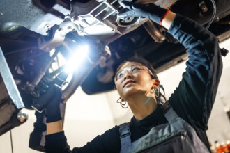 Young woman mechanic wearing safety glasses using flashlight while inspecting car chassis in a