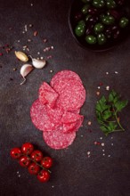 Composition of sliced salami, with fresh tomatoes, garlic and green olives, on a dark surface,