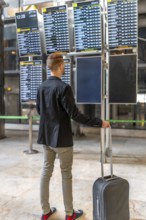 Businessman with trolley suitcase looking at timetable screen while waiting his flight departure at
