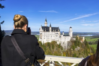 Woman photographing Neuschwanstein Castle in front of a picturesque landscape under a clear sky,