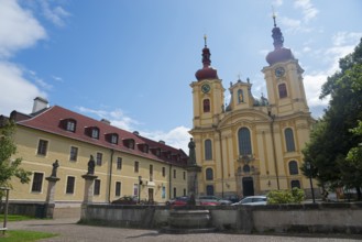 Baroque church next to a building with red roofs and statues, clear blue sky, monastery and