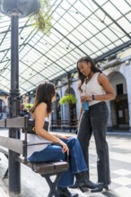 Two female friends chatting and holding reusable coffee cups while standing and sitting on a bench