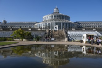 Large historic greenhouse reflected in a pond, palm house with glass facade, blue sky, Botanical