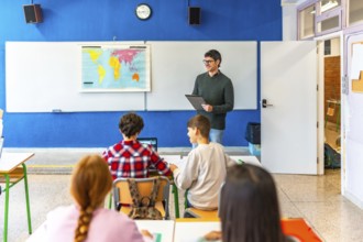 Geography teacher explaining a lesson holding a clipboard and smiling to elementary school students