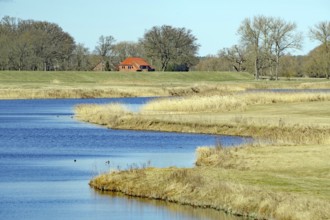 A calm river flows through a green landscape with a red house in the distance under a clear sky,