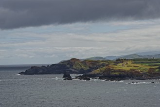A rocky coastline with green hills in the background under a cloudy sky, Fenais da Luz, Sao Miguel