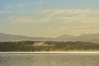 Fog over a calm lake under a clear sky with mountains in the background, Lake Mahinapua, Ruatapu,