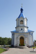 A small white Orthodox church with a blue roof stands in sunny weather against a green backdrop,