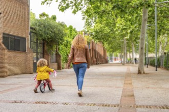 Rear view of a caucasian mother walking aside a girl learning to ride a bicycle in the street