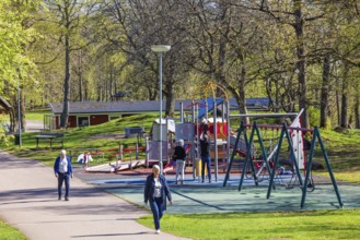 Playground in a public park with children playing and people walking on a footpath at springtime,
