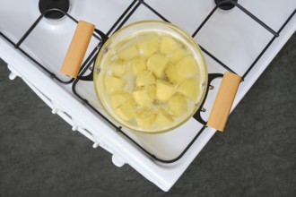 Sliced yellow potatoes are boiling in a clear glass pot on a white stove with wooden handles. The