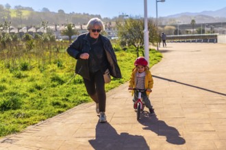 Grandmother jogging behind her grandson riding a balance bike in a park, having fun together on a