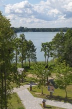 Lake with surrounding trees and well-kept path under a blue sky with clouds, view from the
