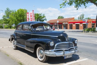 A black vintage car is parked in a car park next to a street with surrounding trees and shops,