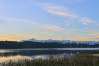 A calm, misty lake in morning sunlight with mountains in the background, Lake Mahinapua, Ruatapu,